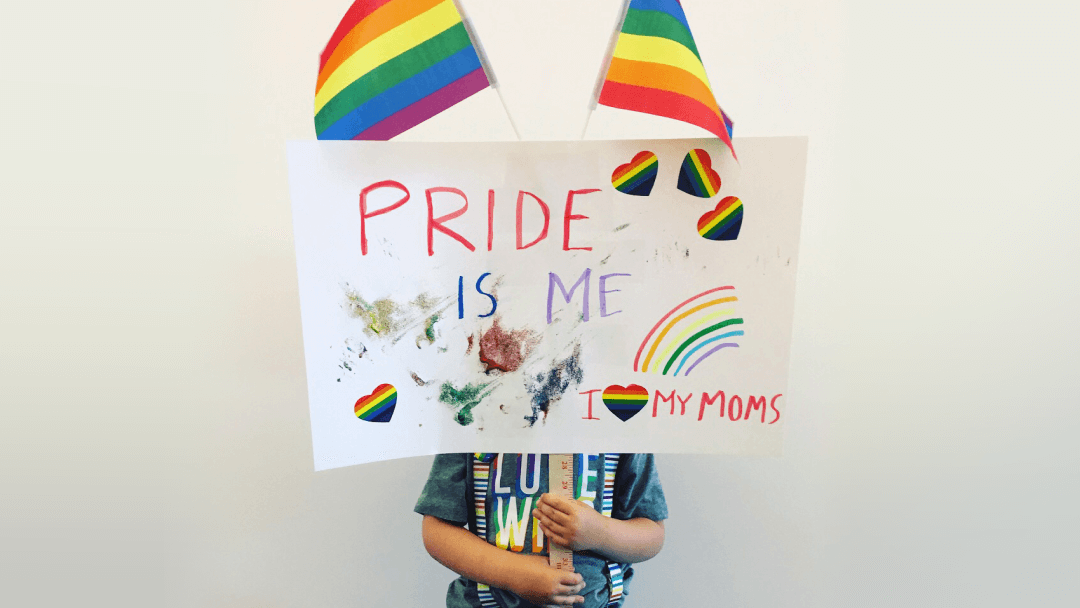 A child holds up a poster with the words 'pride is me' and 'I love my moms' with two rainbow flags attached.